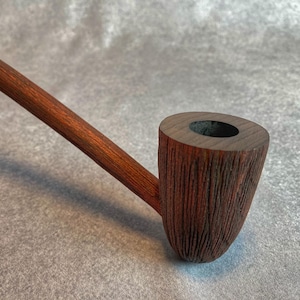 Puede incluir: Una pipa de madera hecha a mano con un acabado marrón oscuro. La pipa presenta un cuenco texturizado y un tallo largo y delgado. El cuenco tiene forma cilíndrica con un interior oscuro. La pipa está diseñada para fumar.