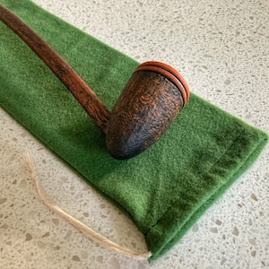 Puede incluir: Una pipa de fumar larga de madera con un acabado marrón y negro. La pipa está apoyada en una bolsa de terciopelo verde.