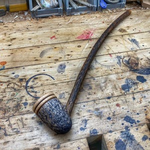 May include: A wooden smoking pipe with a dark brown finish and a bowl made of wood and a light brown material. The pipe is lying on a wooden surface with a lot of scratches and paint splatters.