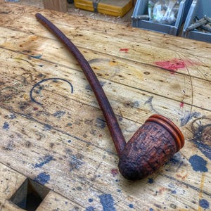 May include: A wooden smoking pipe with a brown and black finish. The pipe has a long, curved stem and a bowl with a textured surface.