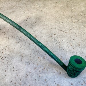 May include: A long, green wooden pipe with a dark green bowl. The pipe is lying on a light gray surface.