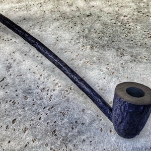 May include: A long, dark wooden pipe with a curved stem and a round bowl. The pipe is lying on a light gray surface.