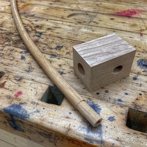 May include: A wooden dowel rod with a slight curve, lying on a worn wooden workbench. A small wooden block with two holes drilled through it sits next to the rod.