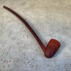 May include: A long, curved wooden pipe with a dark red finish. The pipe has a simple, rounded bowl and a long, slender stem.