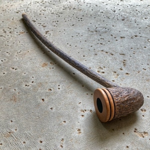 May include: A handcrafted wooden pipe with a dark, curved stem and a textured, rounded bowl. The bowl features a dark opening and is accented with a lighter wood ring. The pipe rests on a textured, light gray surface.
