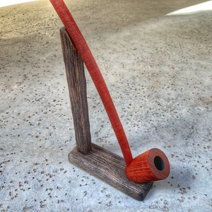 May include: A red wooden pipe resting on a wooden stand. The stand is made of dark wood with a rough texture.