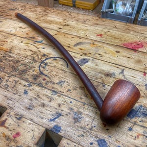 Puede incluir: Una pipa larga de madera marrón oscura con una boquilla curva y una cazoleta redonda. La pipa está apoyada sobre una superficie de madera con muchas marcas.