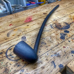 Puede incluir: Una pipa larga de madera negra con una boquilla curva y una cazoleta redonda. La pipa está sobre una superficie de madera.