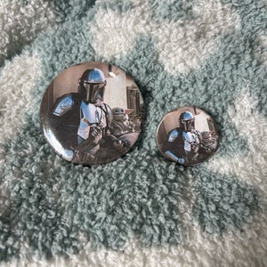 May include: Two round button pins featuring a character from the Star Wars series, The Mandalorian, with a small green alien character. The larger pin is silver with a black background and the smaller pin is silver with a white background.