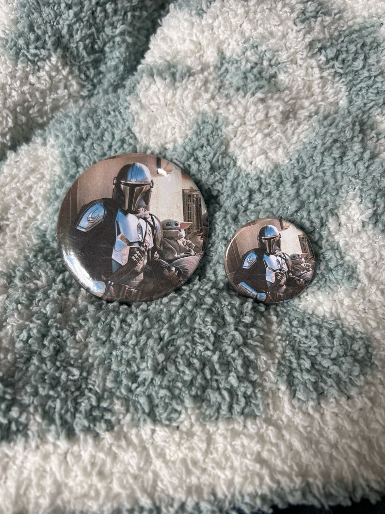 May include: Two round button pins featuring a character from the Star Wars series, The Mandalorian, with a small green alien character. The larger pin is silver with a black background and the smaller pin is silver with a white background.