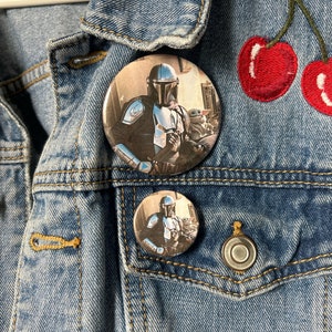 May include: Two round button pins featuring a character from the Star Wars franchise, The Mandalorian, with a small green alien character, Grogu, in his arms. The larger pin is on a denim jacket with a red embroidered cherry design.