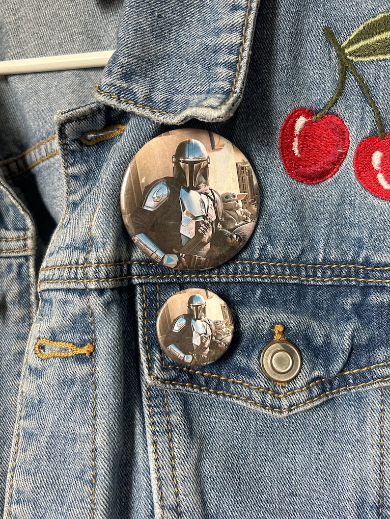 May include: Two round button pins featuring a character from the Star Wars franchise, The Mandalorian, with a small green alien character, Grogu, in his arms. The larger pin is on a denim jacket with a red embroidered cherry design.