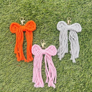 Puede incluir: Tres llaveros de macramé con forma de lazo en naranja, rosa y gris, cada uno con un cierre dorado. Los llaveros están colocados sobre hierba verde. Los lazos tienen un diseño texturizado y anudado con largas colas deshilachadas.