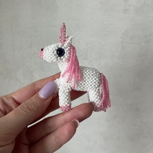 Könnte beinhalten: Ein kleines weißes Perlen-Einhorn mit rosa Mähne und Schwanz. Das Einhorn hat ein blaues Auge und ein rosa Horn.