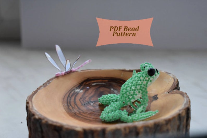 May include: A green beaded frog figurine with black eyes, sitting on a wooden bowl with a pink beaded dragonfly beside it. The image includes the text "PDF Bead Pattern".