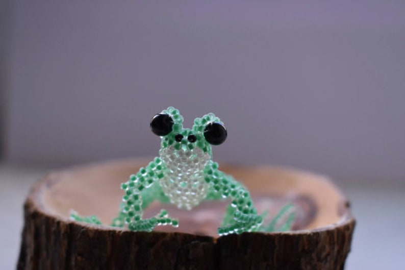 May include: A green and white beaded frog figurine with black eyes, sitting on a wooden log.