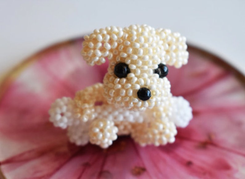 May include: A small, white beaded dog figurine with black eyes. The figurine is sitting on a pink and gold surface.