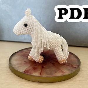 May include: A white beaded horse figurine with black eyes and pink hooves. The horse is standing on a round, brown and pink base.