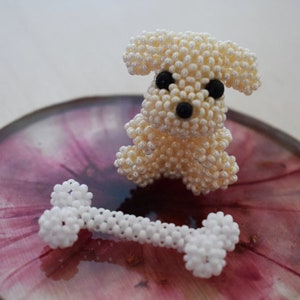 May include: A white beaded dog figurine with black eyes and a black nose, sitting on a pink and white marbled surface with a white beaded bone in front of it.