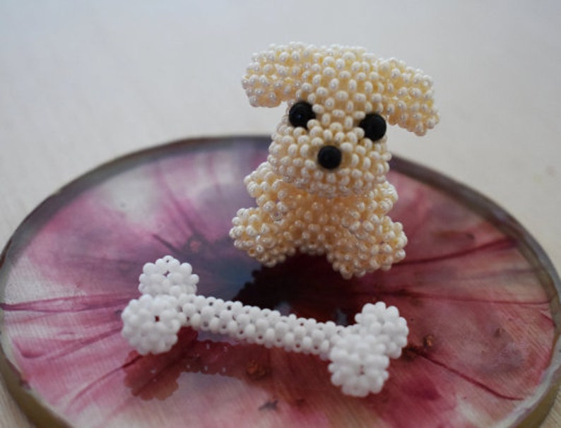 May include: A white beaded dog figurine with black eyes and a black nose, sitting on a pink and white marbled surface with a white beaded bone in front of it.