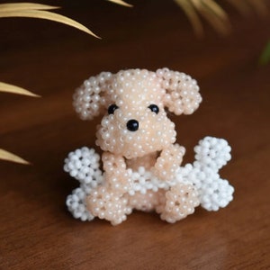 May include: A small, beaded dog figurine with white and light pink beads. The dog is holding two white beaded bones in its paws.