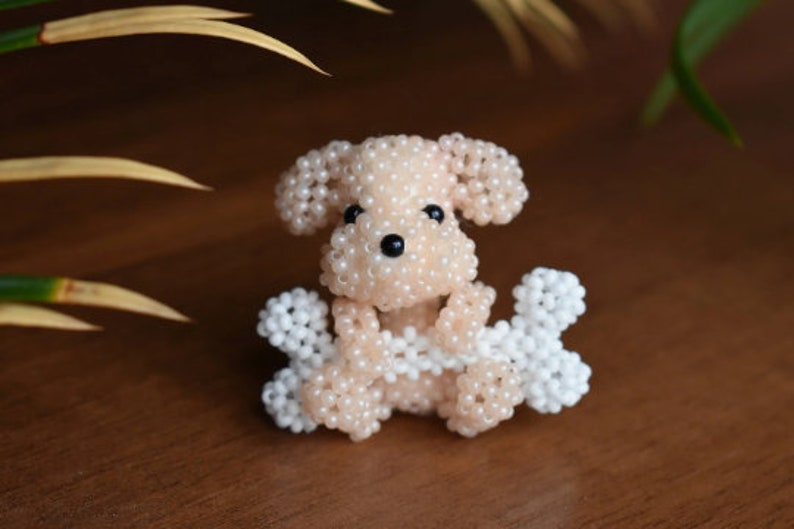 May include: A small, beaded dog figurine with white and light pink beads. The dog is holding two white beaded bones in its paws.