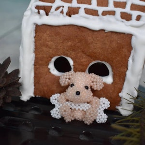 May include: A small, white and pink beaded dog figurine sits in front of a gingerbread house. The house is decorated with white frosting and has a brown roof.