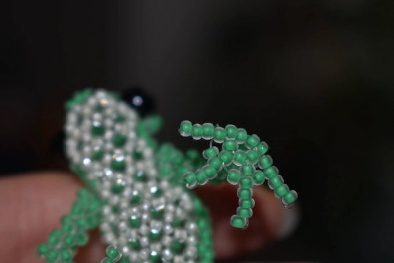 May include: A beaded frog figurine with a green wing-like appendage. The frog is made of white and green beads.