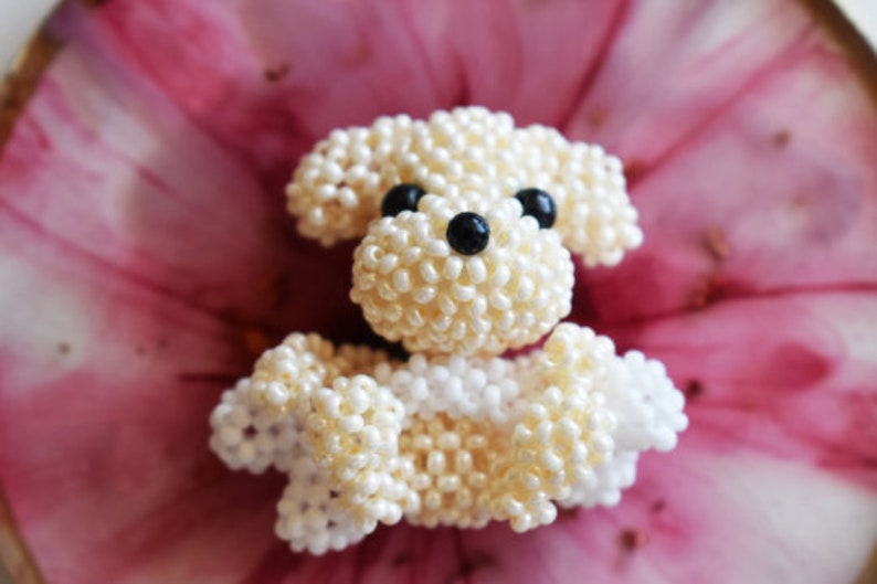 May include: A small, white beaded dog figurine with black eyes. The dog is sitting on a pink and white background.