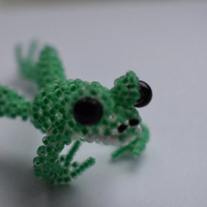 May include: A green beaded frog with black eyes. The frog is sitting on a white surface.
