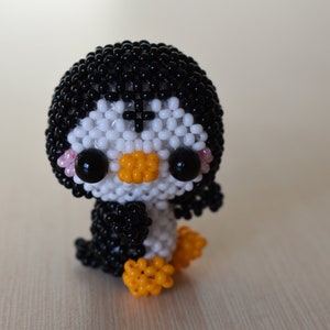 May include: A small, beaded penguin figurine with black and white beads, orange feet, and black eyes. The penguin has a pink blush on its cheeks.