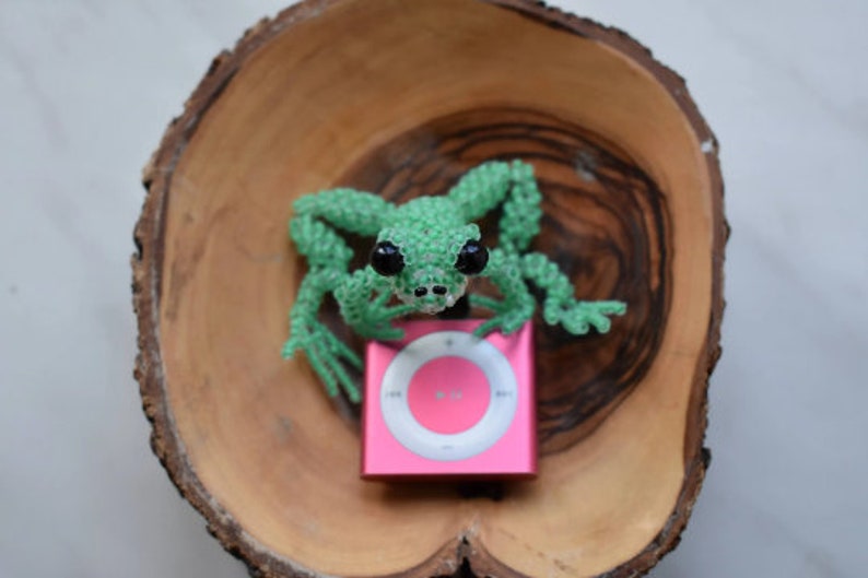 May include: A green beaded frog figurine sits on top of a pink iPod with a white face and a circular button in the center.