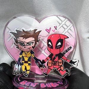 Puede incluir: Un soporte acrílico transparente con ilustraciones de dibujos animados de Wolverine y Deadpool tomados de la mano, sobre un fondo de corazón rosa. La base dice "Best Buds" en letras rosas. Los personajes están con sus trajes característicos.