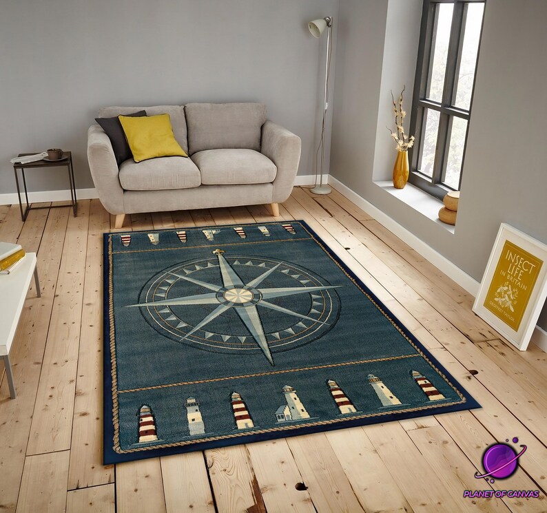 May include: A rectangular blue nautical rug with a compass rose design. The rug has a border of lighthouses and rope detailing. It is placed on a wooden floor in a living room. The rug measures approximately 200cm x 300cm.