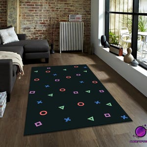 Puede incluir: Alfombra rectangular negra con formas geométricas coloridas, incluyendo círculos, cuadrados, triángulos y cruces. La alfombra está colocada sobre un suelo de madera en un salón. La alfombra tiene un diseño lúdico.