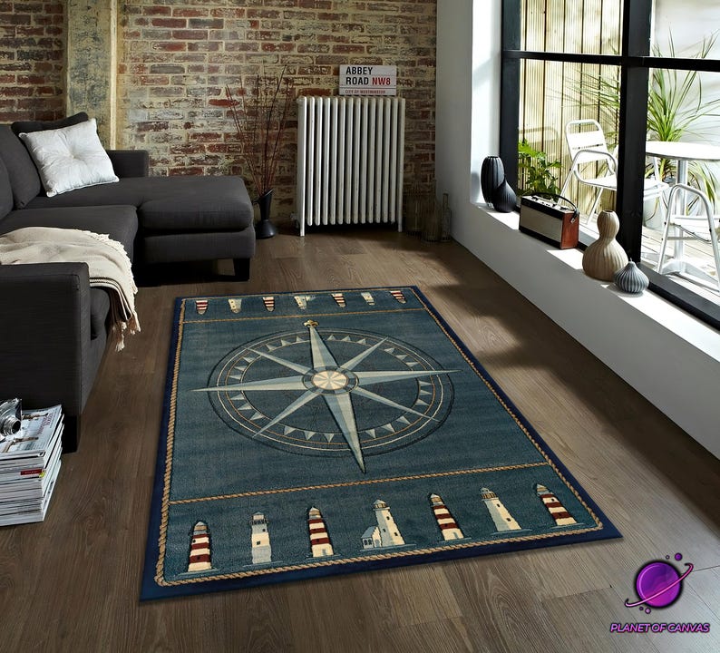 May include: A rectangular blue nautical rug with a central compass rose design. The rug is bordered with lighthouse motifs and rope detailing. The rug is displayed in a living room setting, with a dark sofa.