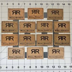 May include: Thirteen rectangular tan labels with a wrinkled texture, each featuring the black logo RR and the text REAL RANCH WEAR. The labels are arranged on a white cutting mat with a grid marked in inches, showing each label is 2 inches wide.