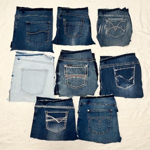 May include: An assortment of folded denim jeans in various shades of blue. Each pair features unique pocket designs, including decorative stitching and embellishments. The jeans are arranged on a neutral background, showcasing the different styles and washes.