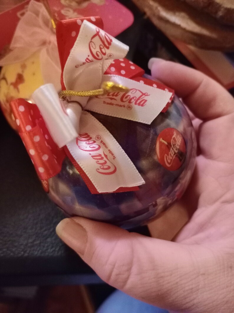 K&ouml;nnte beinhalten: Ein transparenter Weihnachtsbaumschmuck aus Glas mit glitzerndem Galaxie-Design. Er hat ein rot-wei&szlig;es Band mit wei&szlig;en Punkten und ein wei&szlig;es Etikett mit dem Text "Coca-Cola Trade-mark (R)".