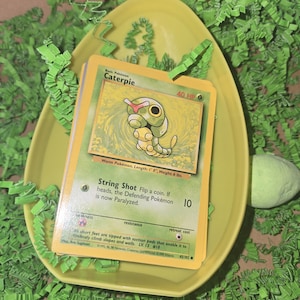 Puede incluir: Cartas coleccionables de Pokémon con el personaje Caterpie, presentadas en un plato amarillo en forma de huevo. Las cartas están rodeadas de hierba de papel verde y un huevo amarillo. La carta Caterpie tiene un diseño verde y amarillo.