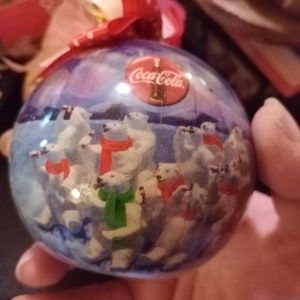 K&ouml;nnte beinhalten: Ein transparenter Weihnachtsbaumschmuck aus Glas mit einer blauen und wei&szlig;en Winterlandschaft mit Eisb&auml;ren und dem Coca-Cola-Logo. Der Schmuck ist mit einem roten Band mit wei&szlig;en Punkten verziert.