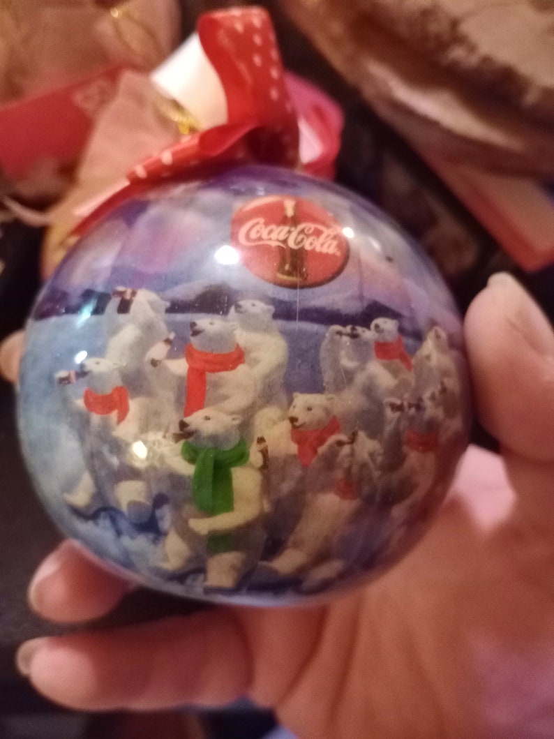 K&ouml;nnte beinhalten: Ein transparenter Weihnachtsbaumschmuck aus Glas mit einer blauen und wei&szlig;en Winterlandschaft mit Eisb&auml;ren und dem Coca-Cola-Logo. Der Schmuck ist mit einem roten Band mit wei&szlig;en Punkten verziert.