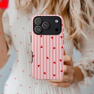Puede incluir: Funda de teléfono a rayas rosas y blancas con pequeños corazones rojos. La funda tiene un módulo de cámara negro y se sostiene en una mano. La funda está diseñada para un teléfono inteligente. Tema romántico.