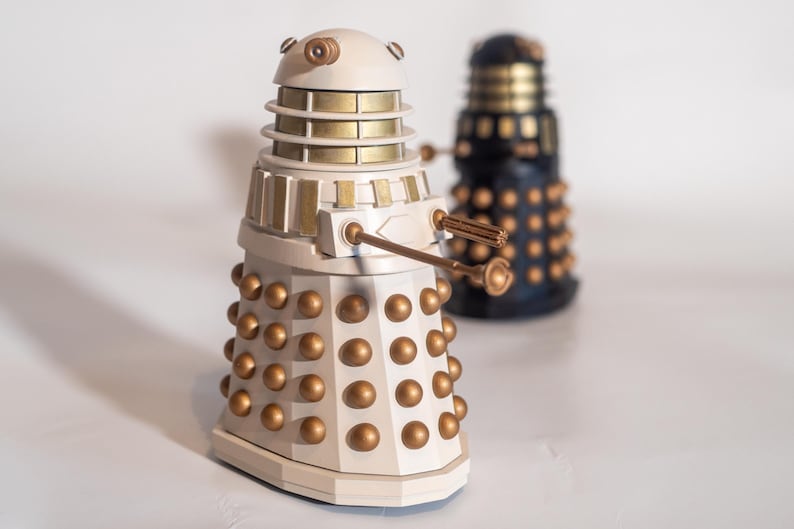 May include: Two Dalek figurines, one white with gold accents and one black with gold accents. Both Daleks have a dome-shaped head with a single eye and a circular base with multiple segments.