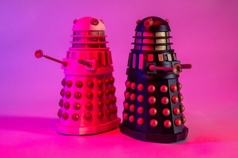 May include: Two Dalek figurines, one white and one black, with gold accents. The Daleks are standing on a pink surface.