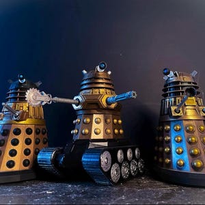 Könnte beinhalten: Drei detaillierte Dalek-Modelle. Die Daleks sind hauptsächlich gold- und braunfarben mit schwarzen Akzenten. Der mittlere Dalek ist auf einem Panzerketten-Sockel montiert und hat eine blaue Kanone. Die anderen beiden Daleks haben verschiedene Armaufsätze.