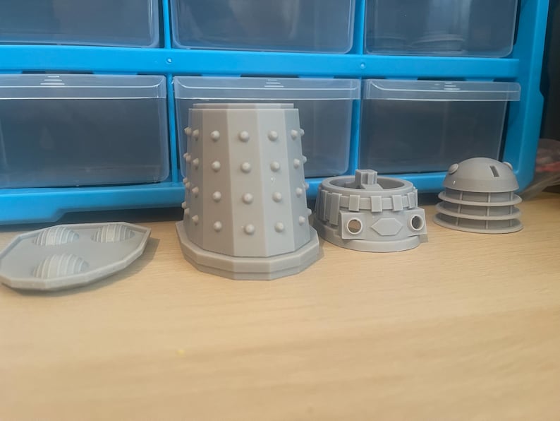 May include: Four gray 3D printed pieces of a Dalek model, including the base, dome, and two sections of the body.
