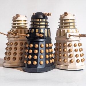May include: Three Dalek figurines, one black and two white, with gold accents. The Daleks are standing on a white surface.