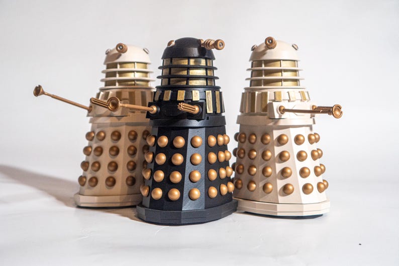 May include: Three Dalek figurines, one black and two white, with gold accents. The Daleks are standing on a white surface.