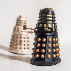 May include: Two Dalek figurines, one black and gold, the other white and gold. The black Dalek is in focus and has a raised arm with a weapon. Both Daleks have a dome-shaped head with a single eye.